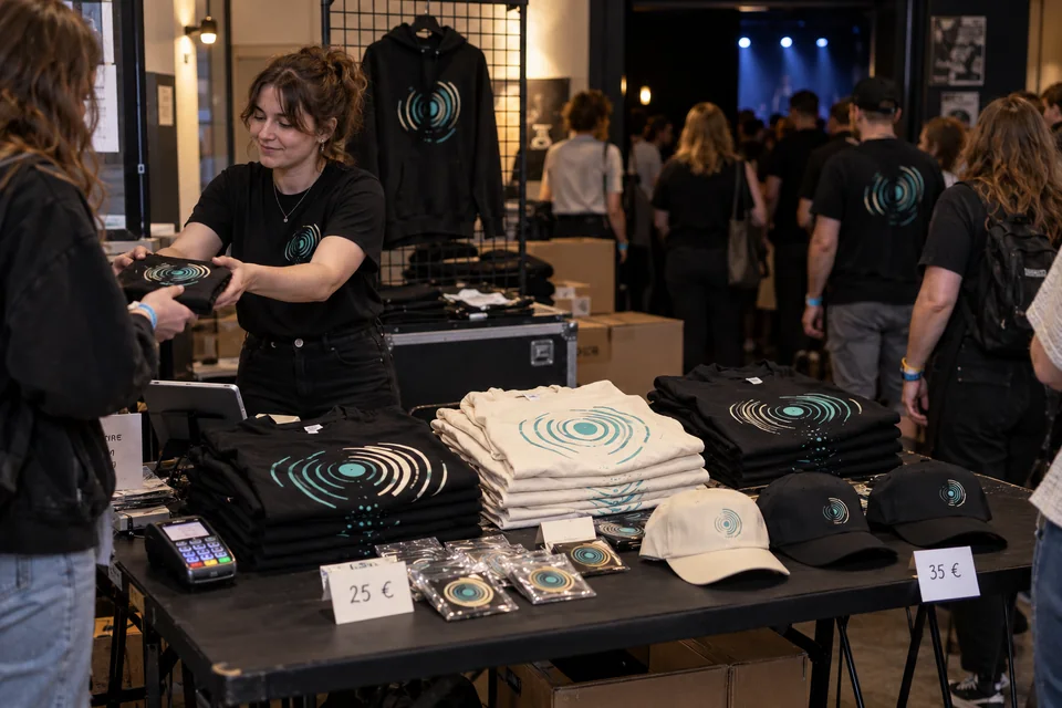 Merch-Verkaufsstand mit bedruckten Shirts, Hoodies, Caps und Stickern im Foyer eines Liveevents.