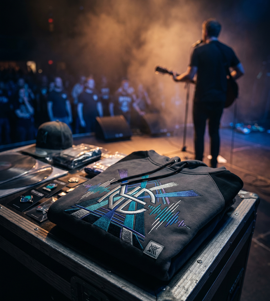 Premium Band Merch und Künstler Merch mit hochwertig besticktem Hoodie auf einem Flightcase vor Konzertbühne mit Musiker, Bühnenlicht, Rauch und After-Show-Atmosphäre