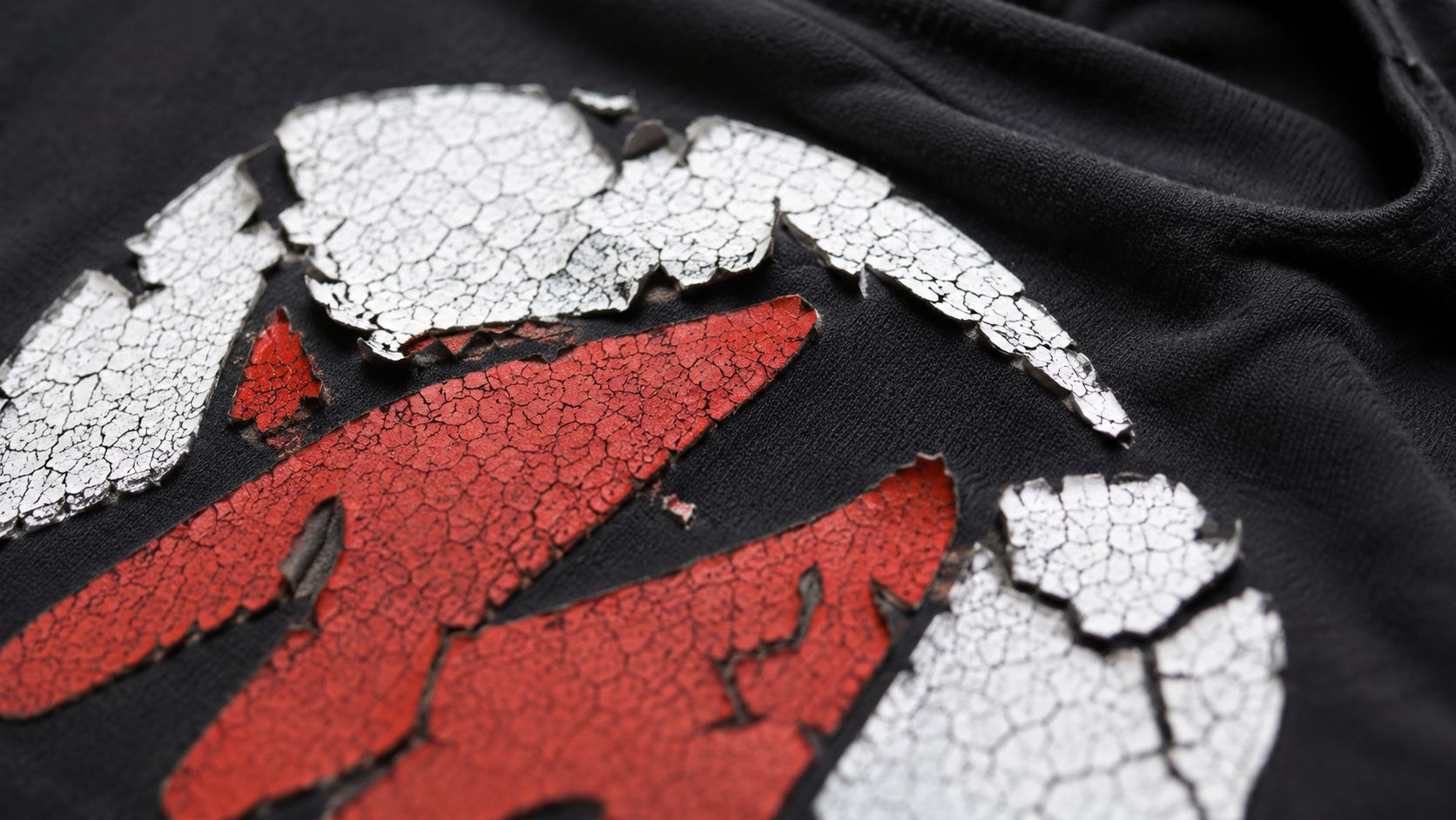 Ein schwarzes T-Shirt mit einem stark rissigen, abgenutzten silbernen und roten Logo-Druck auf dem Stoff.